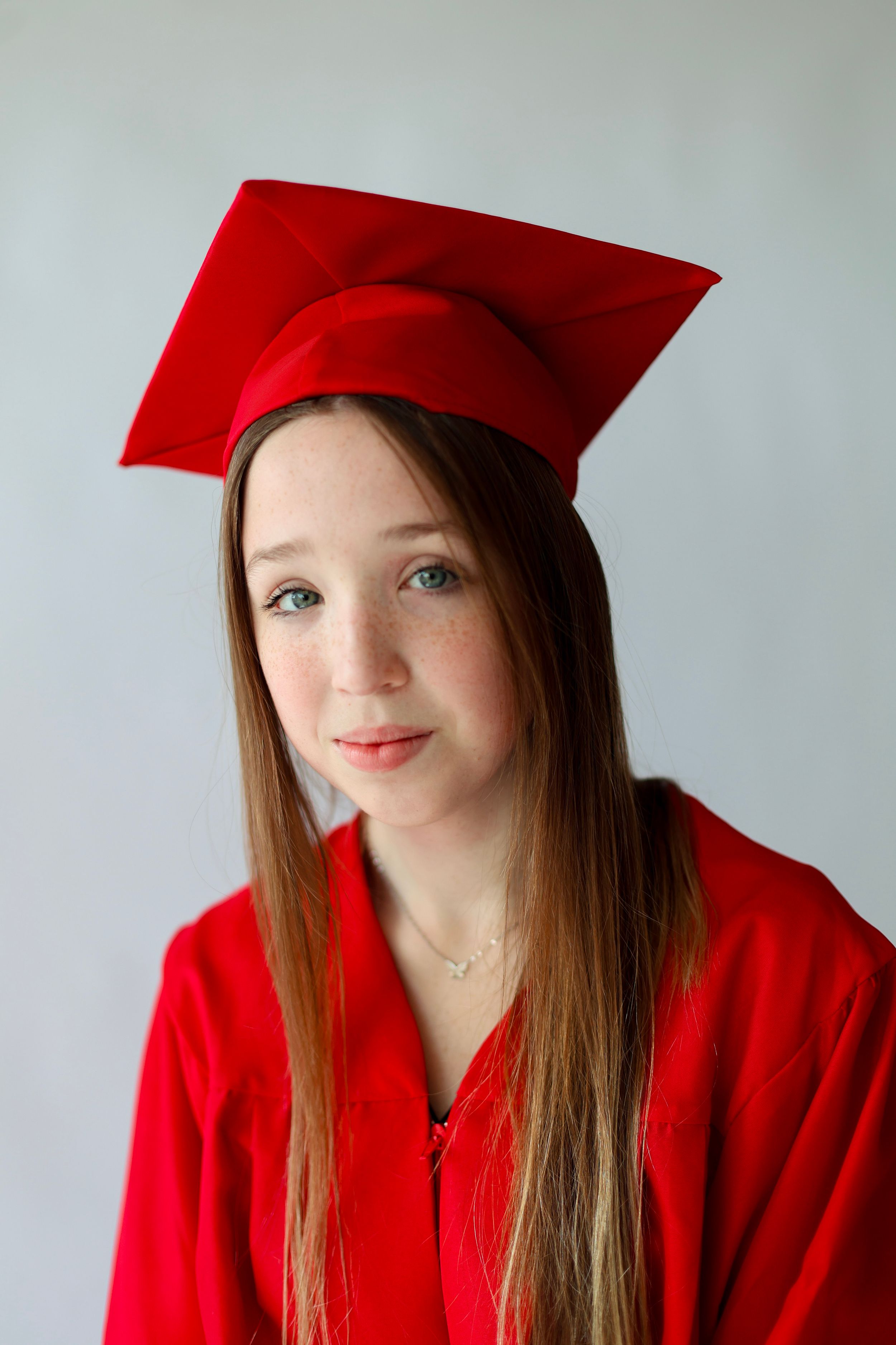 Young girl in red graduation cap and gown smiling for a portrait photo against a light gray background.