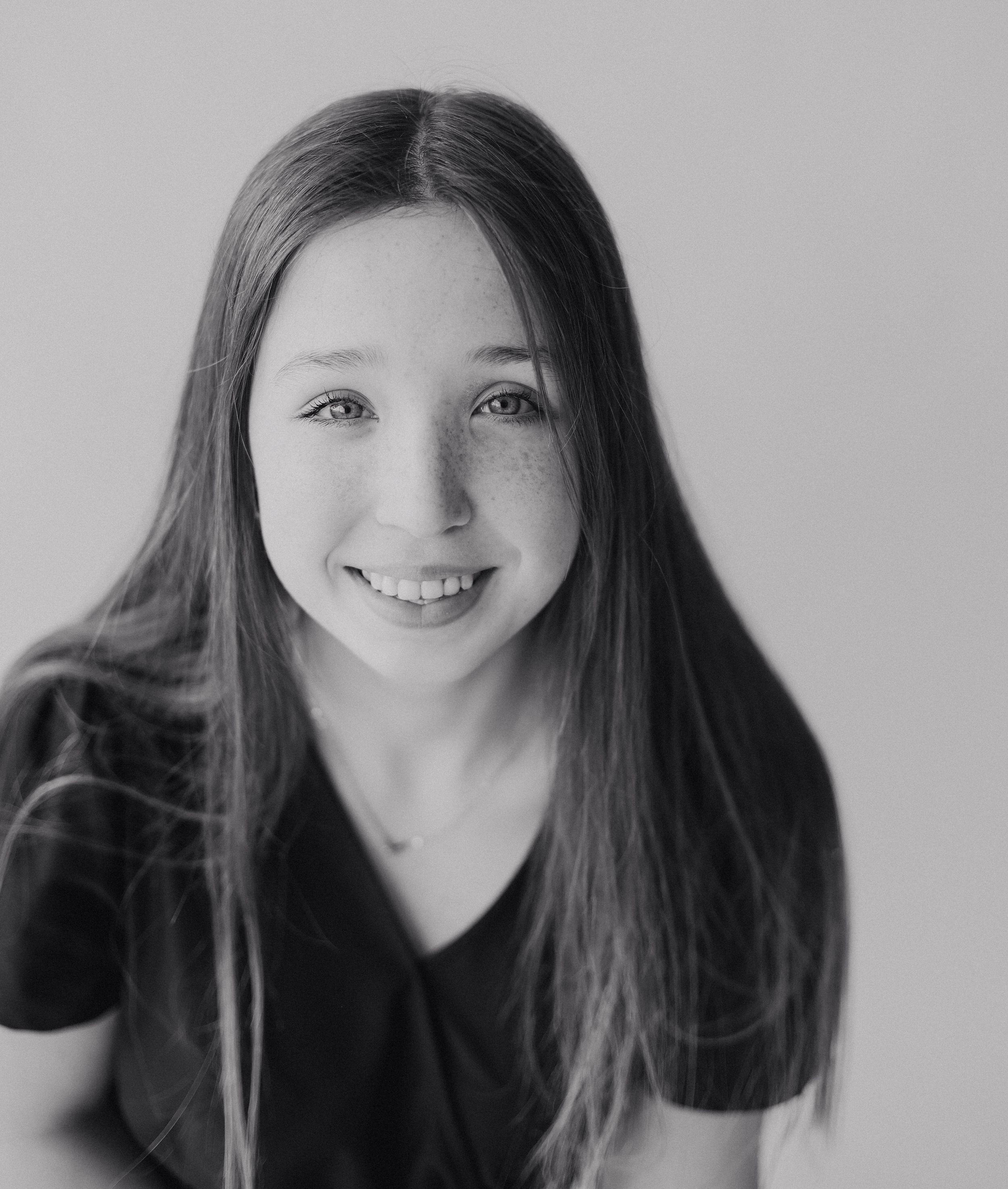 Black and white portrait of a smiling young girl with long hair and freckles against a light background.