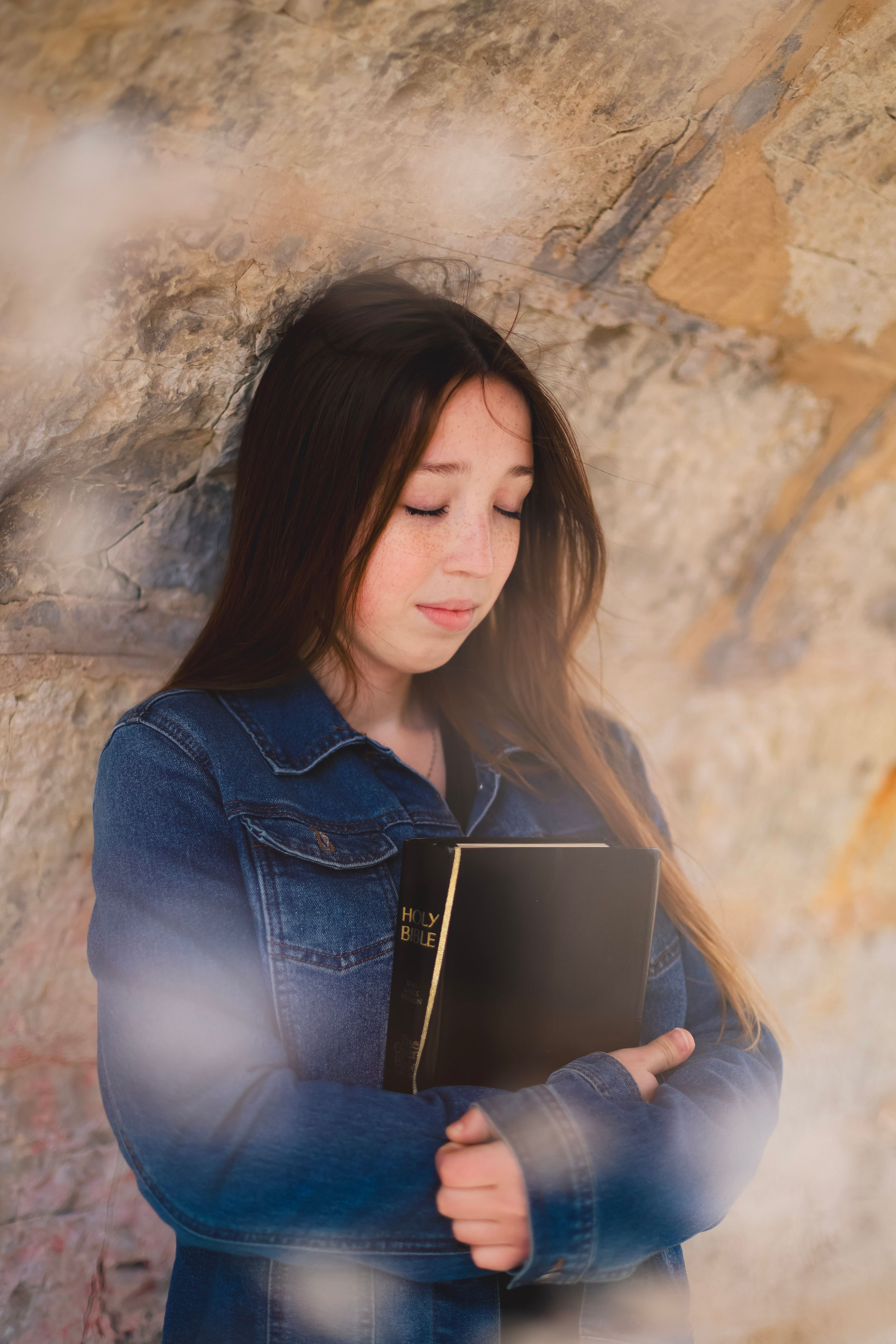 Young girl in denim jacket holding a Holy Bible against a rustic stone wall with soft light effects.