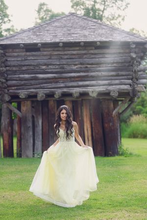 Bride in yellow gown poses on green lawn before rustic log cabin backdrop.