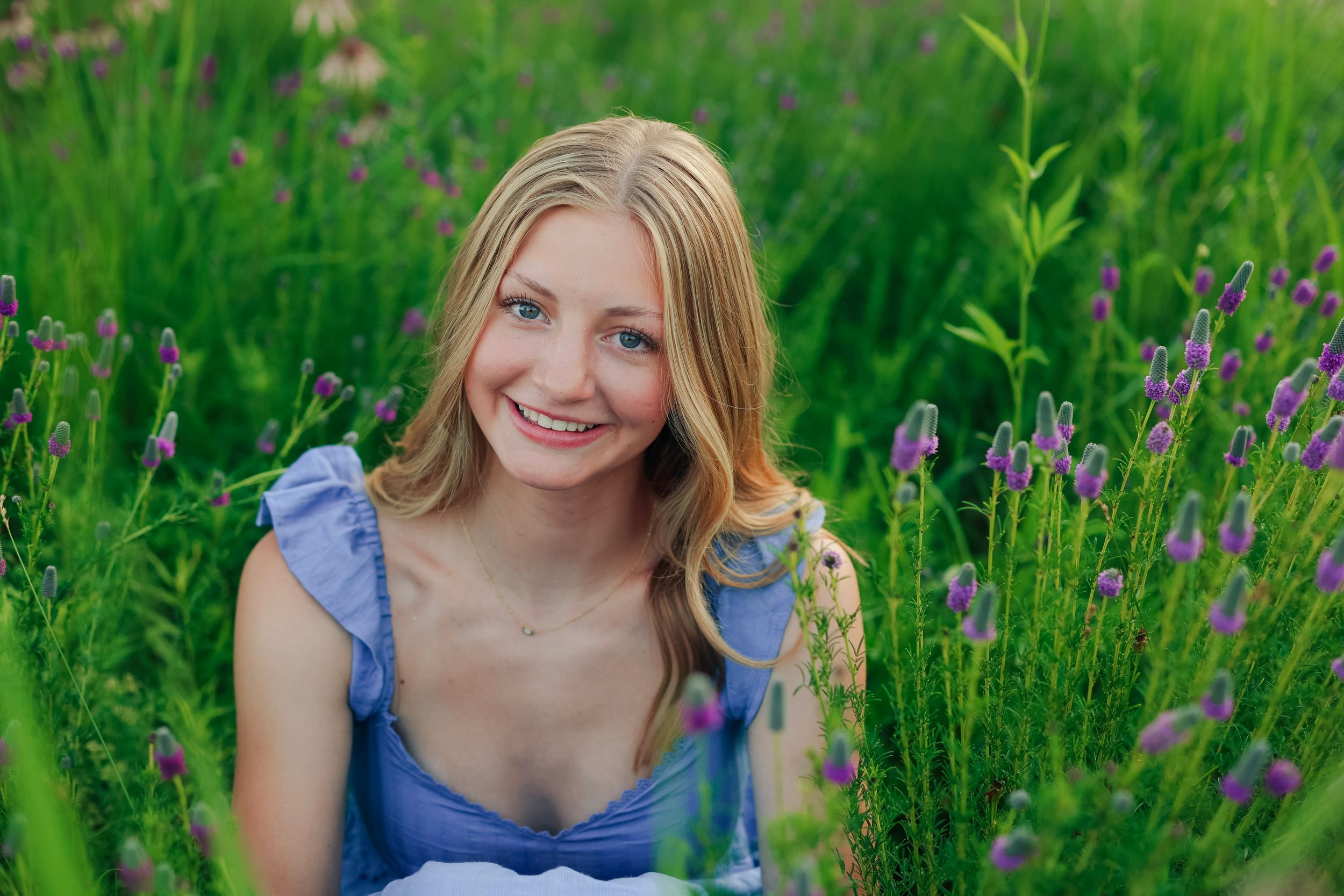 Smiling young blonde woman in a blue dress sitting among vibrant green grass and purple lavender flowers outdoors.