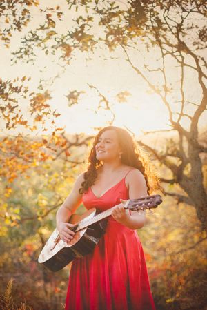 Woman in red dress plays acoustic guitar outdoors during golden hour among autumn trees.