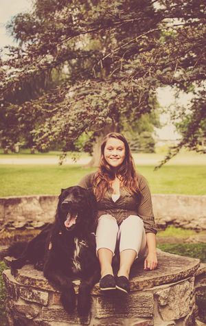 Smiling young woman sitting outdoors on stone wall with large black dog beside her in green park.