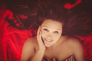 Smiling woman with flowing dark hair lying on red fabric, hand on cheek, looking up with blue eyes.