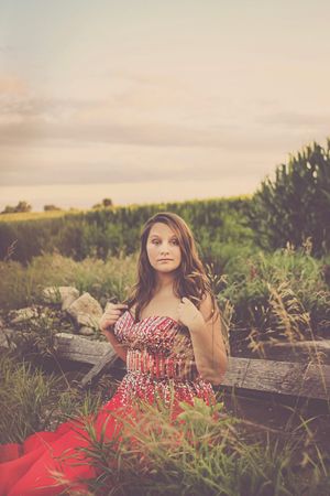 Young woman in red floral dress sitting by rustic wooden fence in a golden field at sunset.