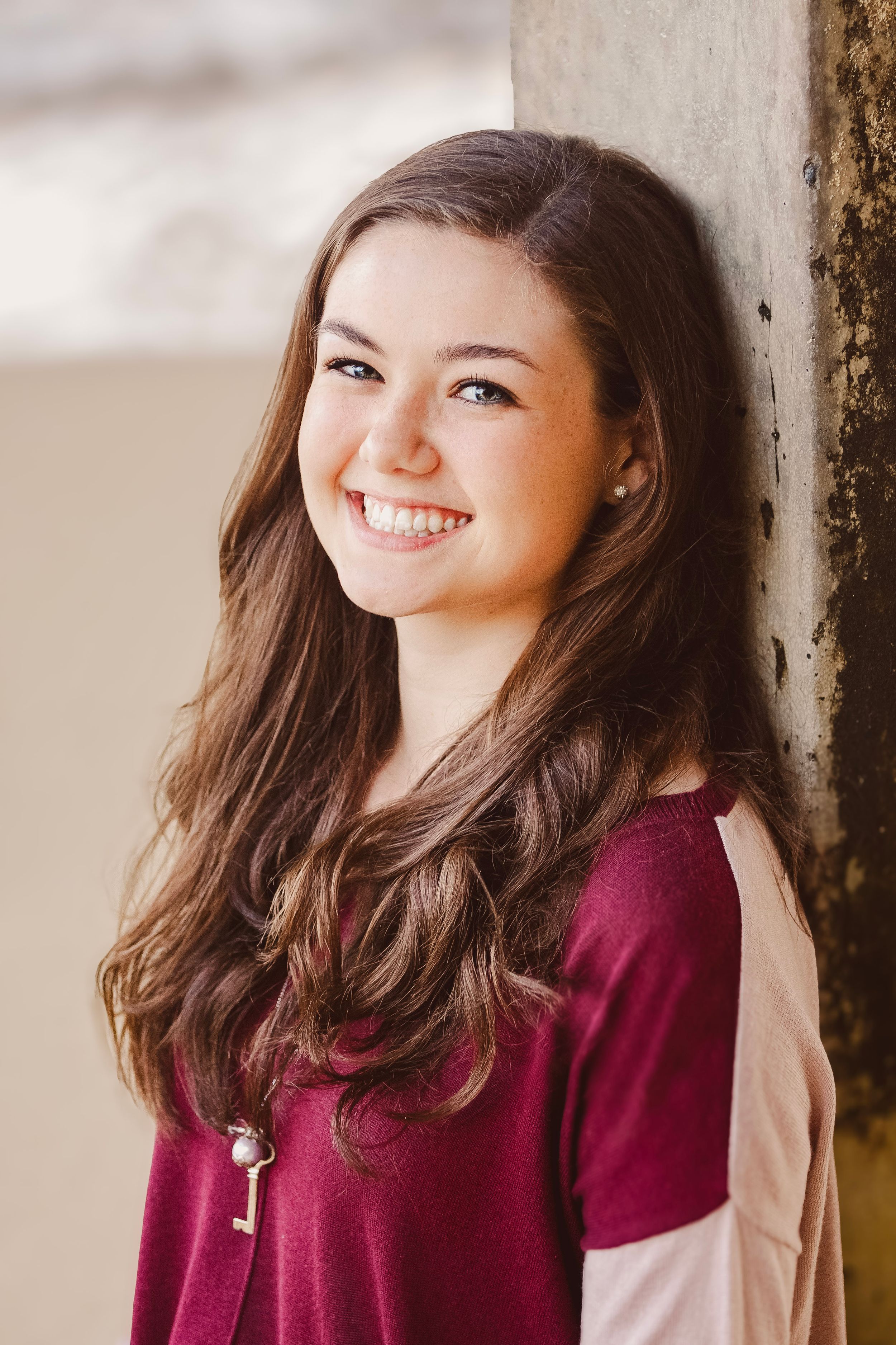 Smiling young woman with long brown hair wearing a burgundy top, leaning against a pillar outdoors.