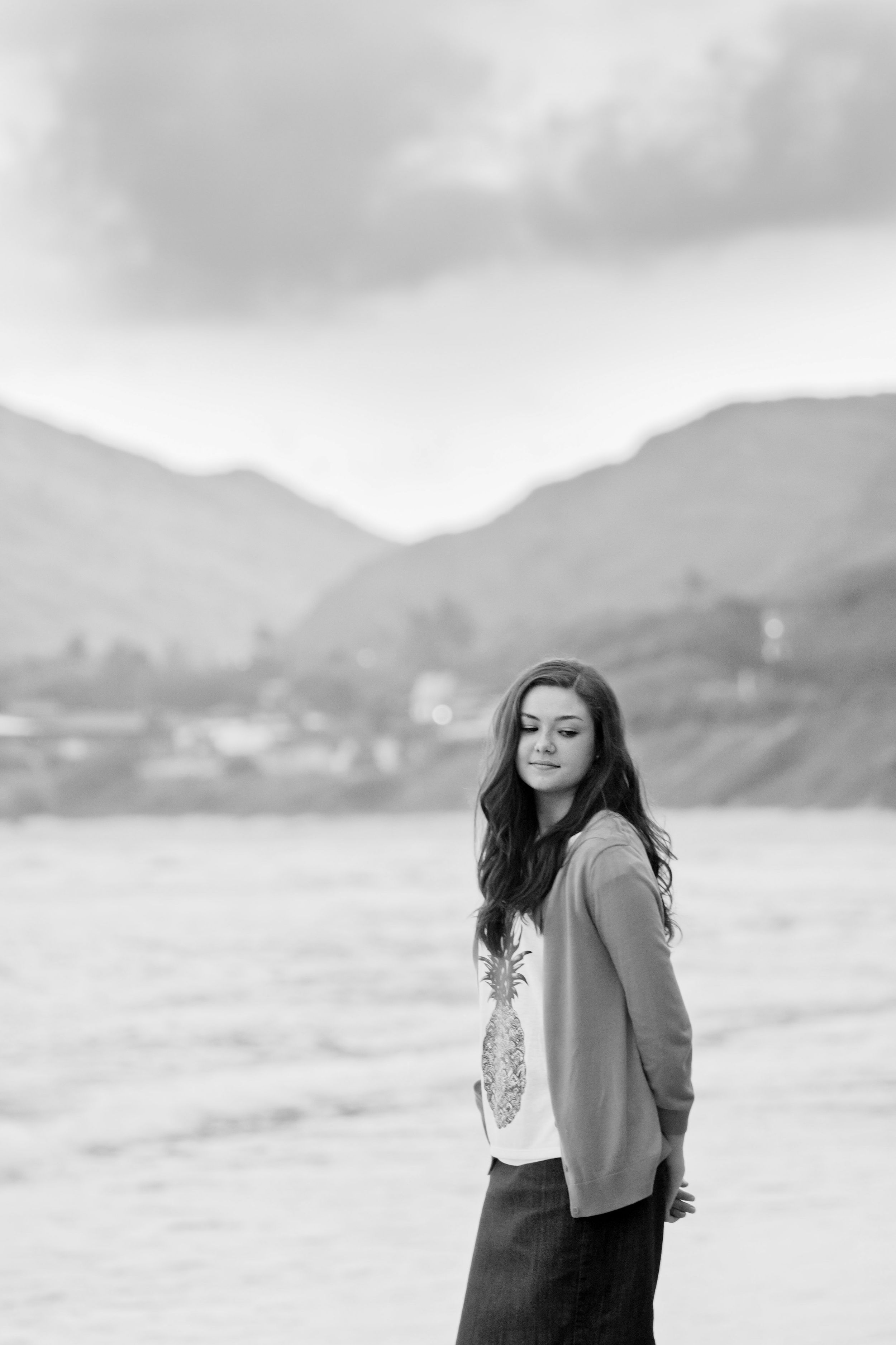 Young woman with long hair stands on a beach in black and white, mountains visible in the background.