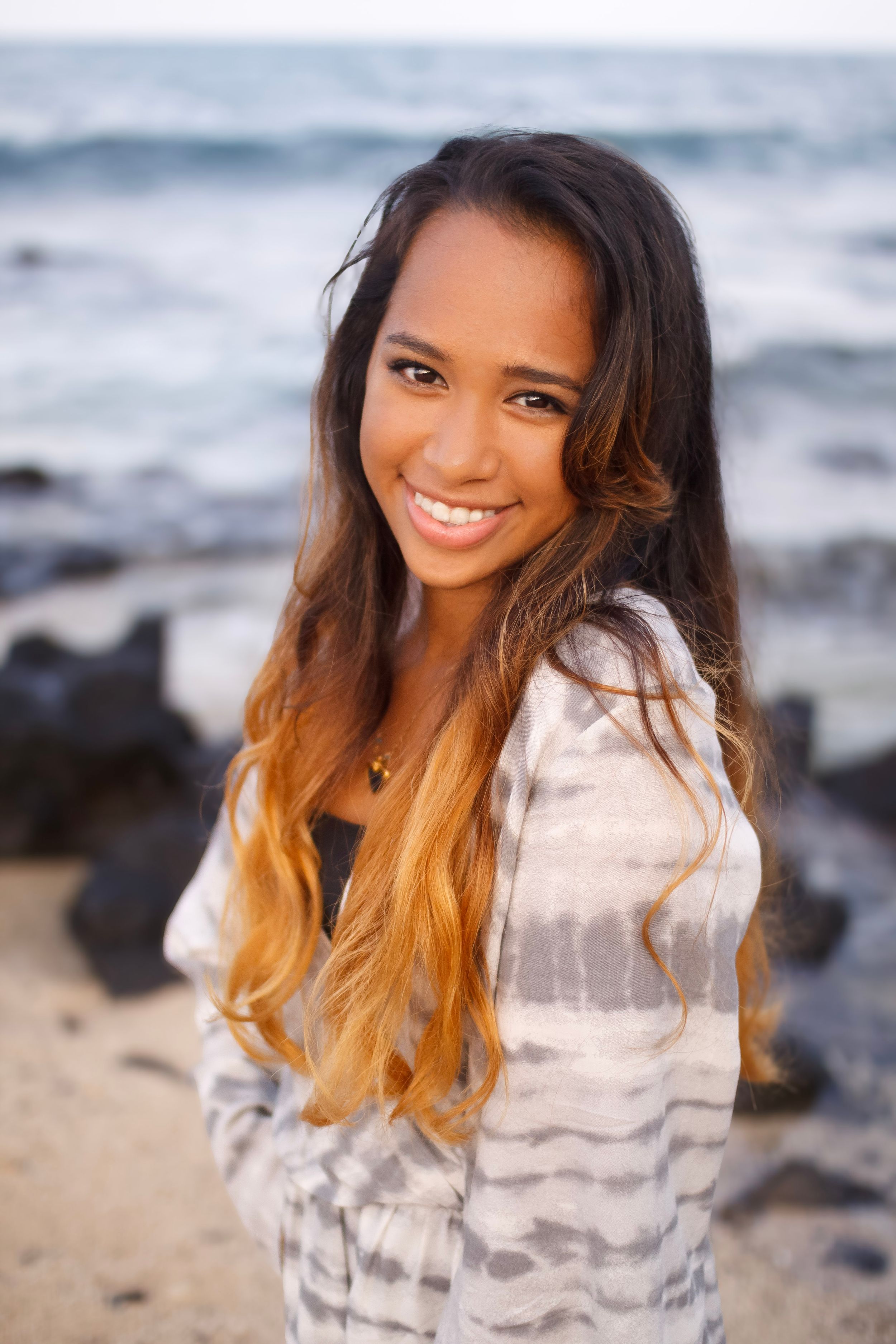 Young woman with ombre hair smiling at beach with rocky shoreline and ocean waves in background.