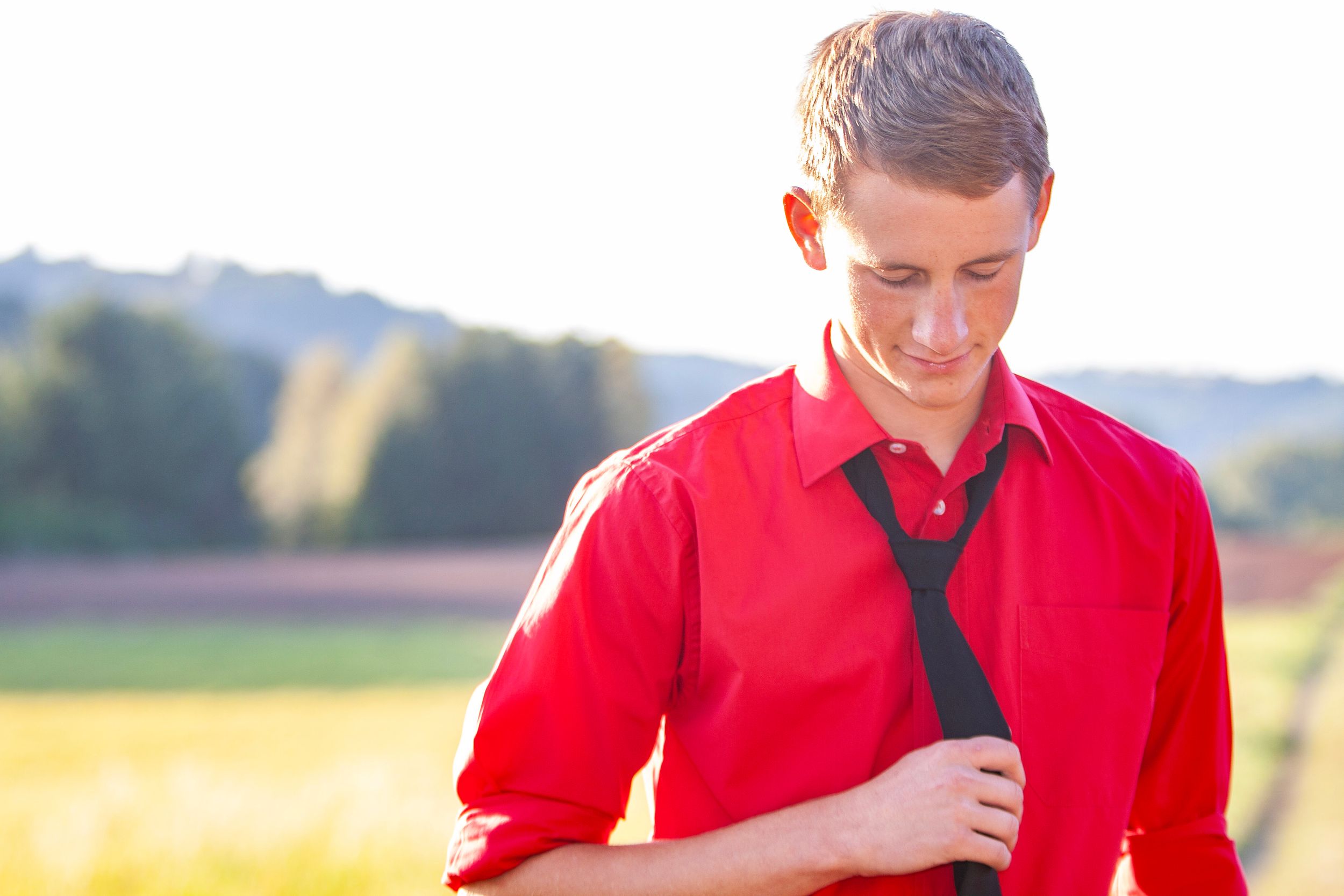 Young man in red shirt adjusting black tie outdoors in a sunny field.
