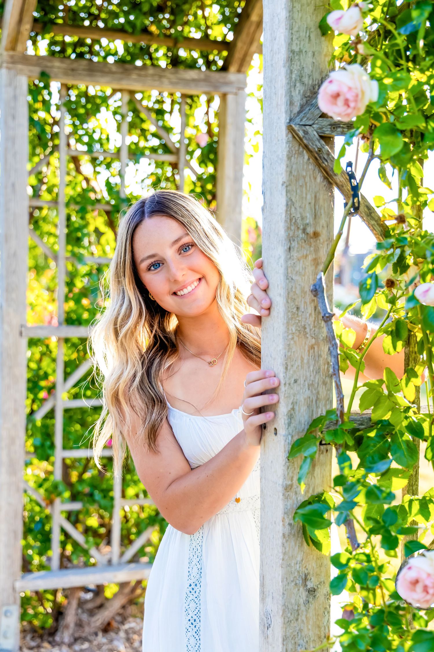 Young blonde woman in white dress smiling beside a wooden garden trellis with pink climbing roses.
