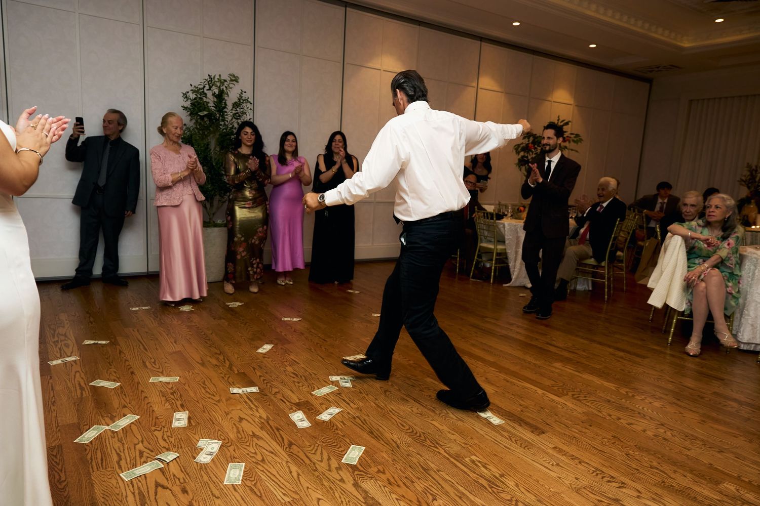 Man dancing at wedding reception with money on floor, guests clapping and watching in elegant ballroom.