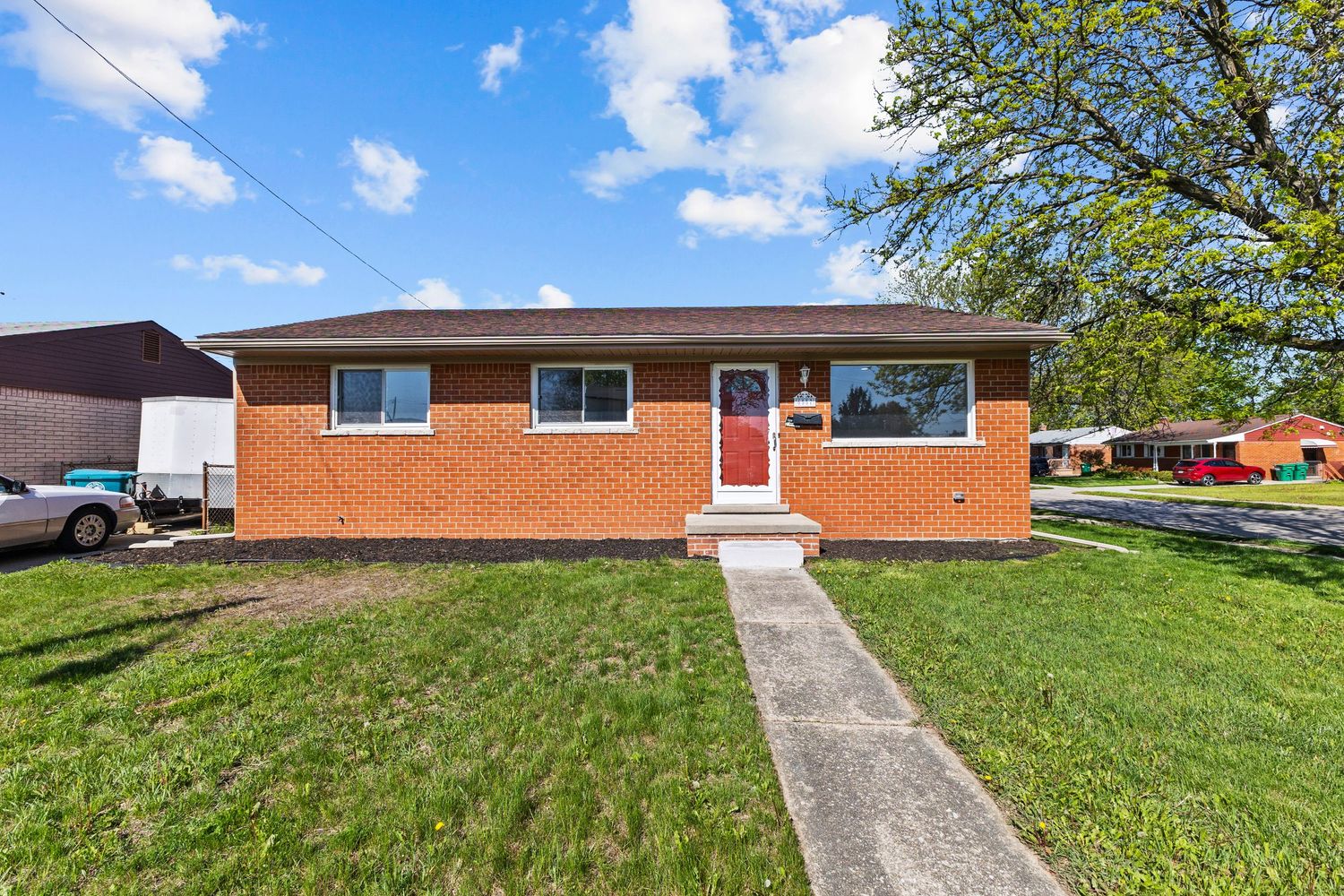 Brick ranch-style home with red front door, green lawn, and mature trees under a blue sky.
