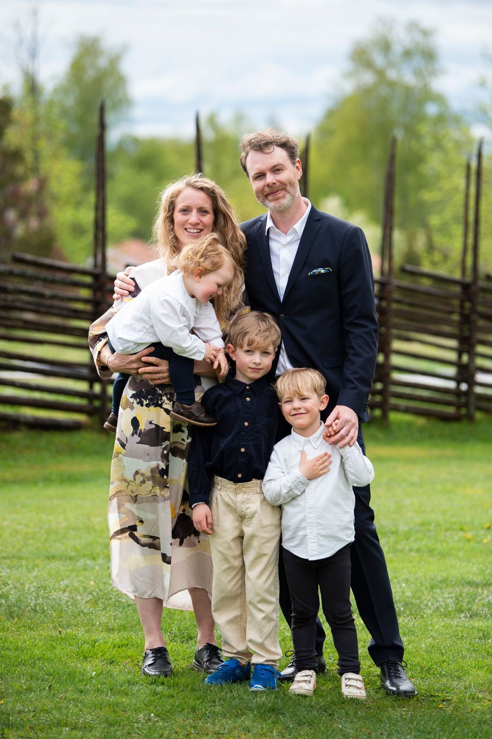 Happy family of five posing outdoors on green grass, parents with three young boys dressed in formal attire.