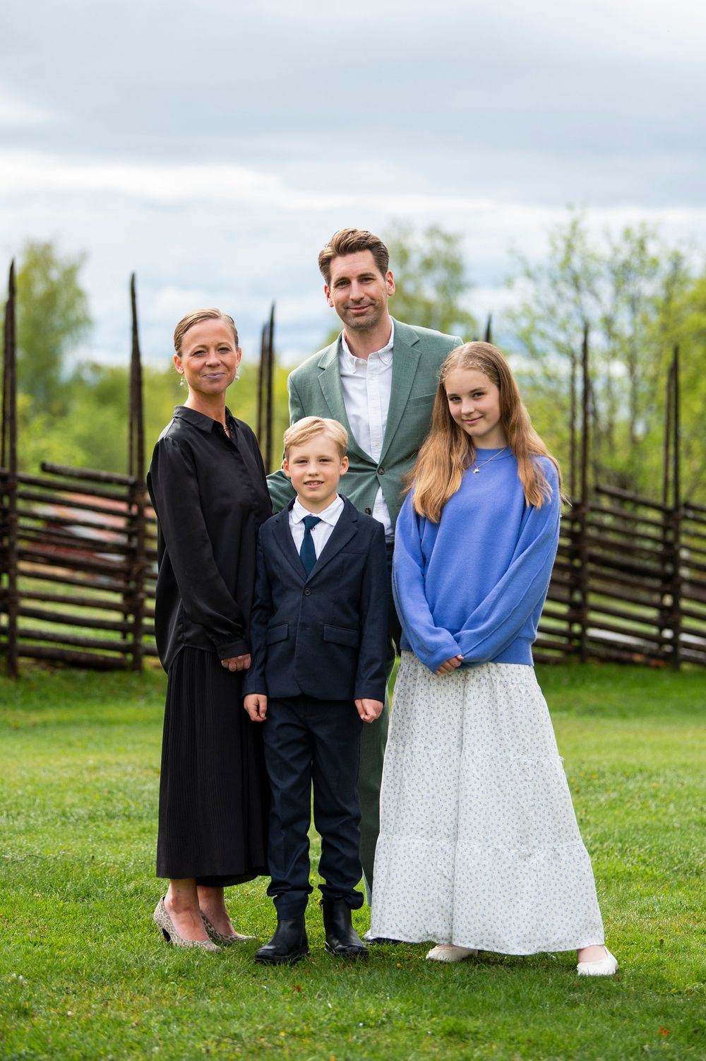 Family of four posing outdoors on green grass with a wooden fence and trees in the background.