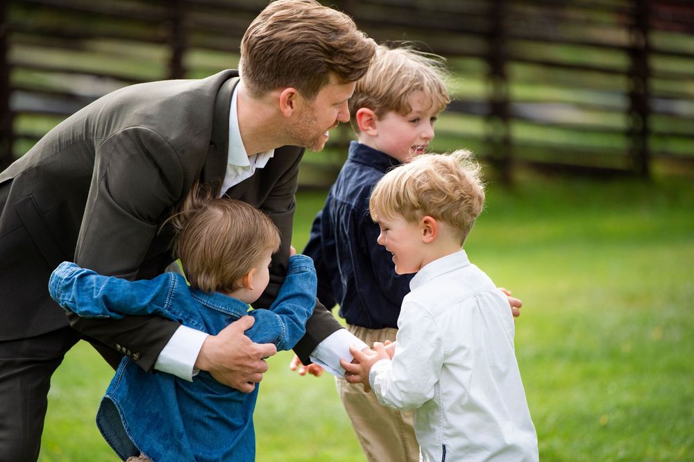 Man in suit bending down to interact with three young boys on a green lawn outdoors.