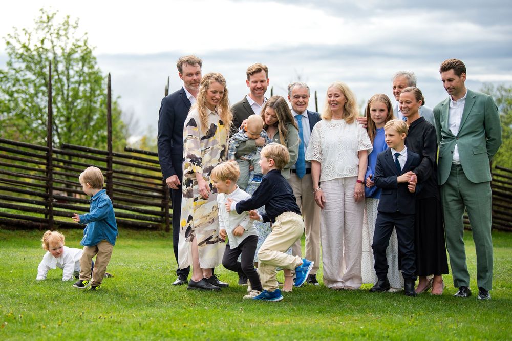 Extended family gathering outside on lawn, adults posing with young children playing during a special occasion.