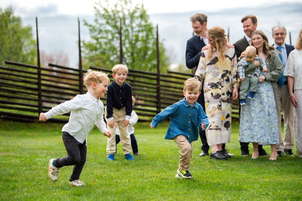 Children playing and running on green grass while adults watch and smile in an outdoor garden setting.