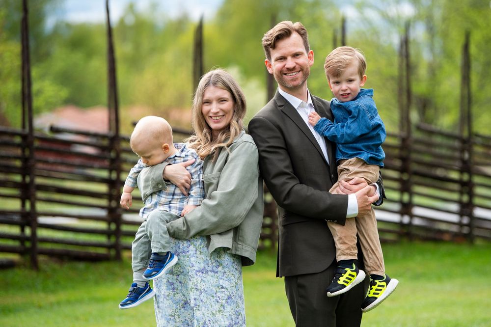 A smiling family of four poses outdoors, with parents each holding a young toddler boy in a green countryside setting.