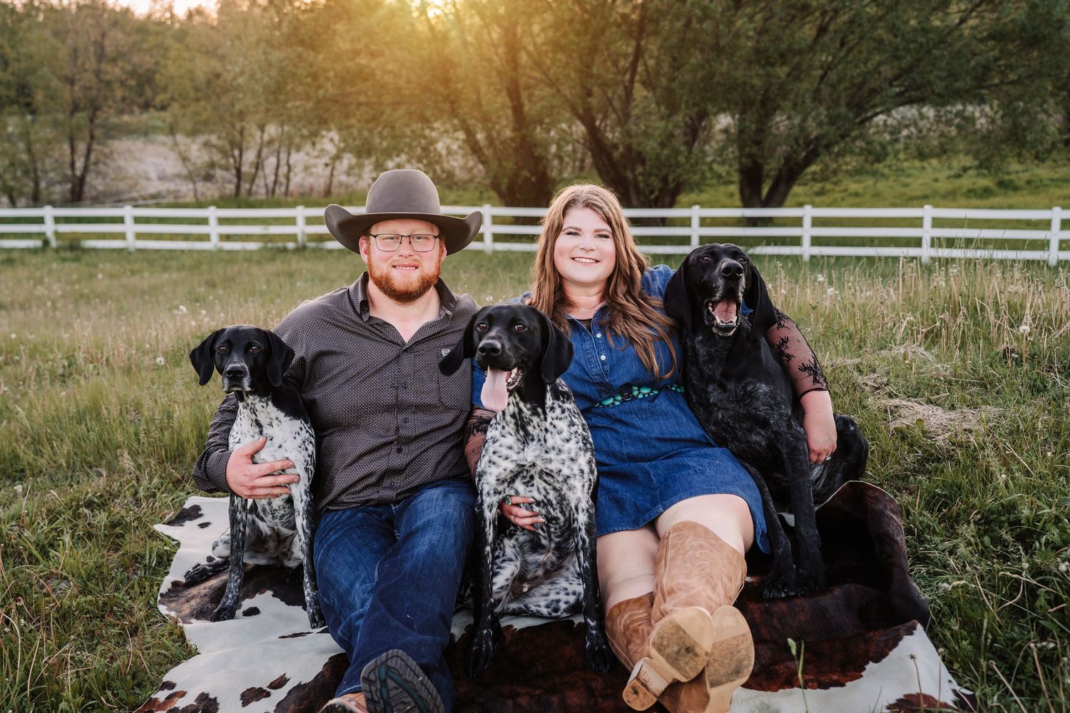 Couple sitting on cowhide rug in field at sunset with three black dogs, white fence and golden trees in background.