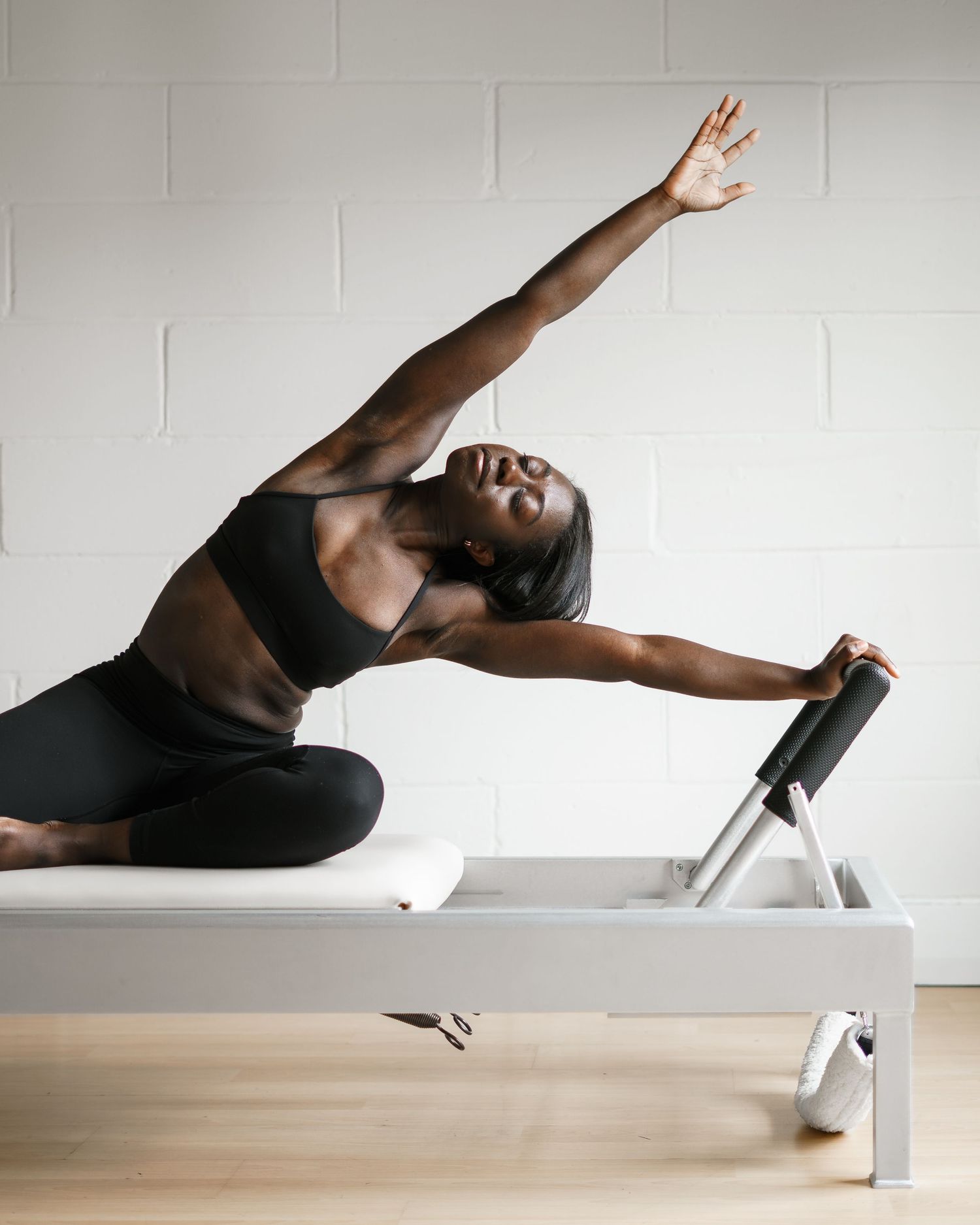 Woman performing a side stretch exercise on a Pilates reformer machine in a bright studio with white paneled walls.