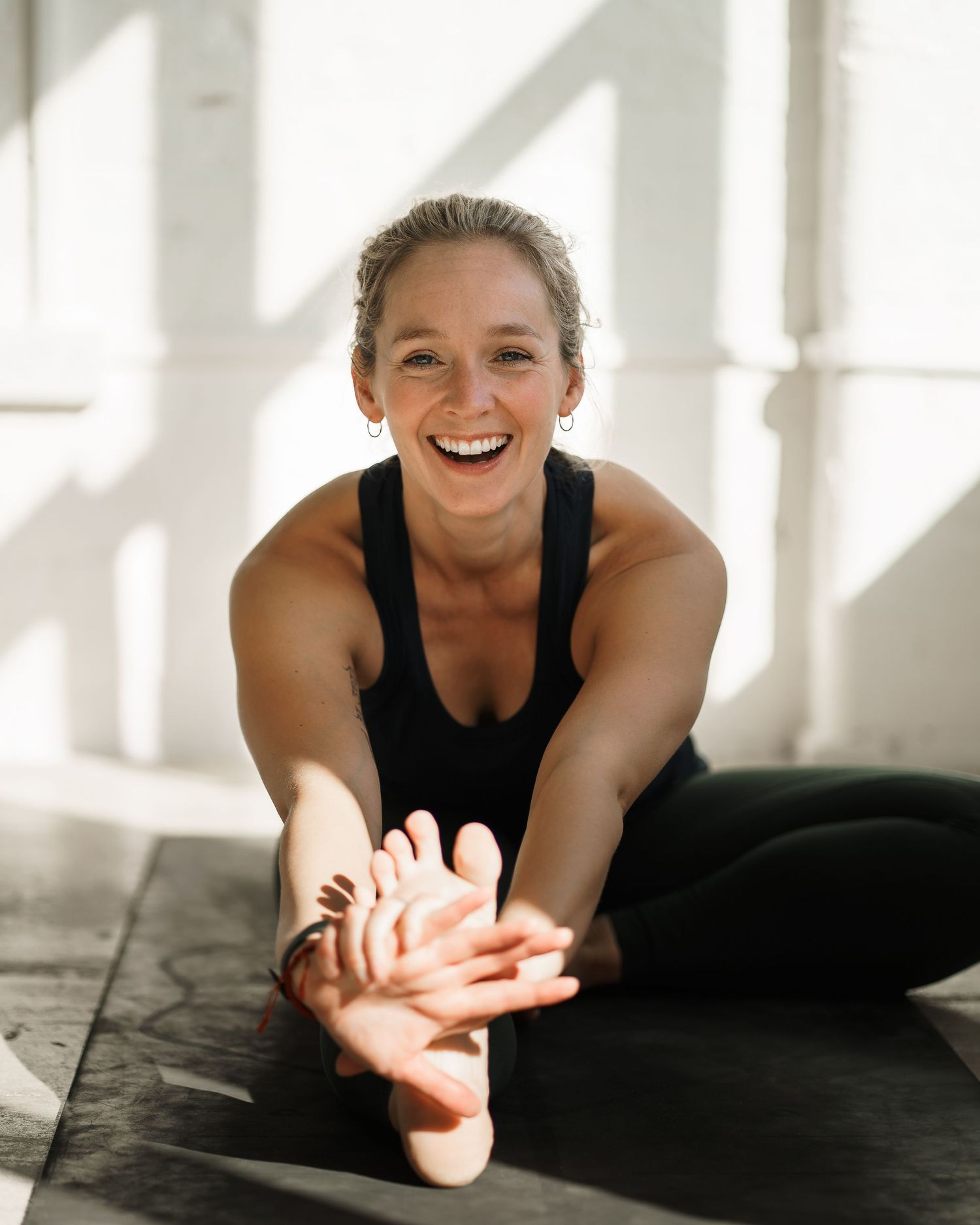Smiling woman in black athletic wear stretching on yoga mat in bright studio, reaching toward feet.