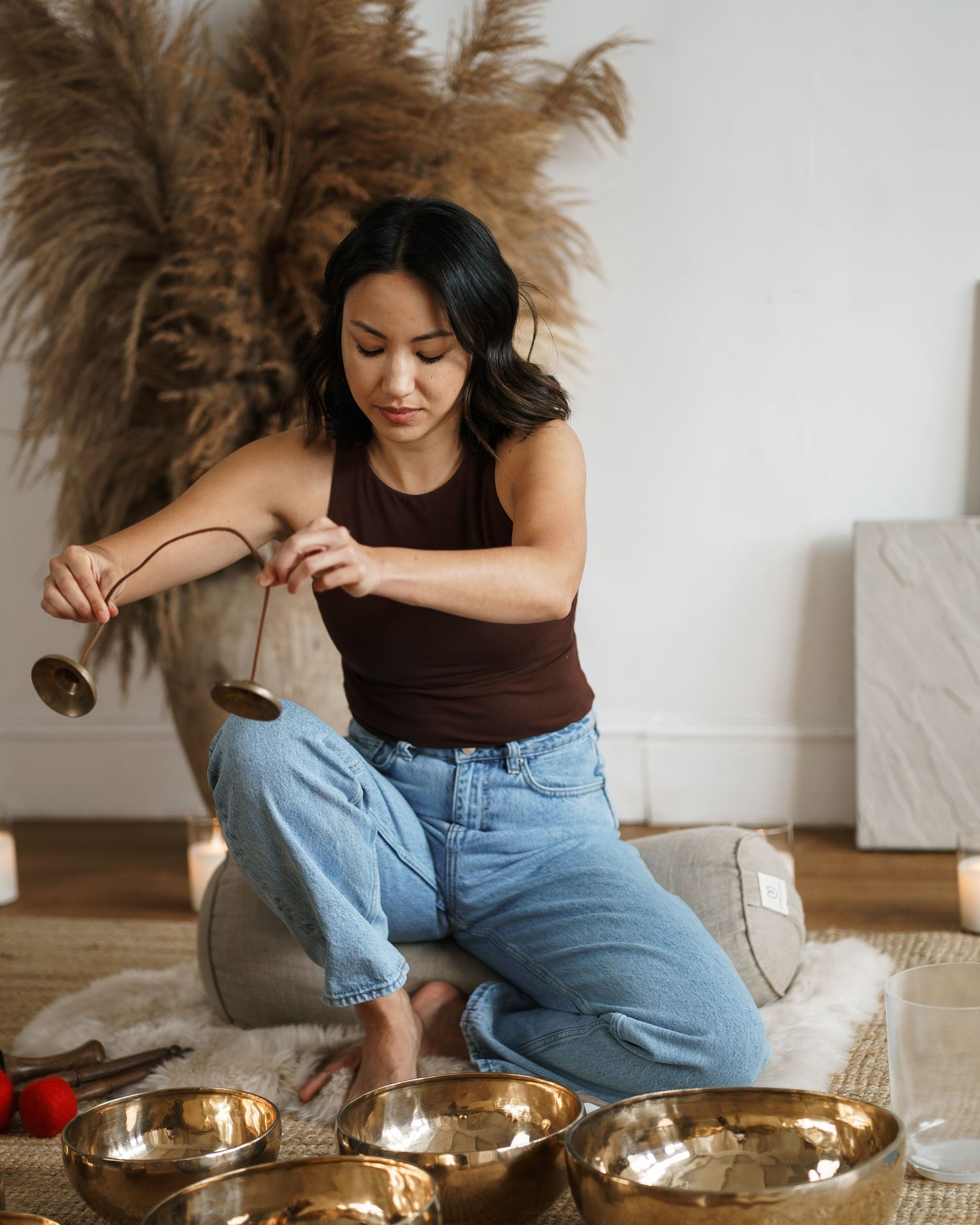 Woman playing Tibetan singing bowls while kneeling on a rug in a serene studio with pampas grass decor.