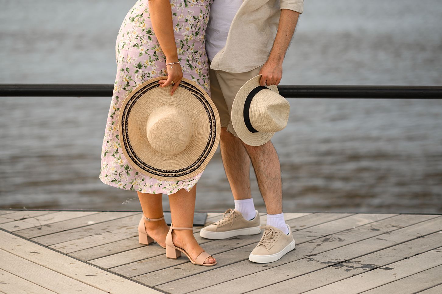 Couple standing on wooden pier holding straw hats, wearing summer outfits, with water in background.
