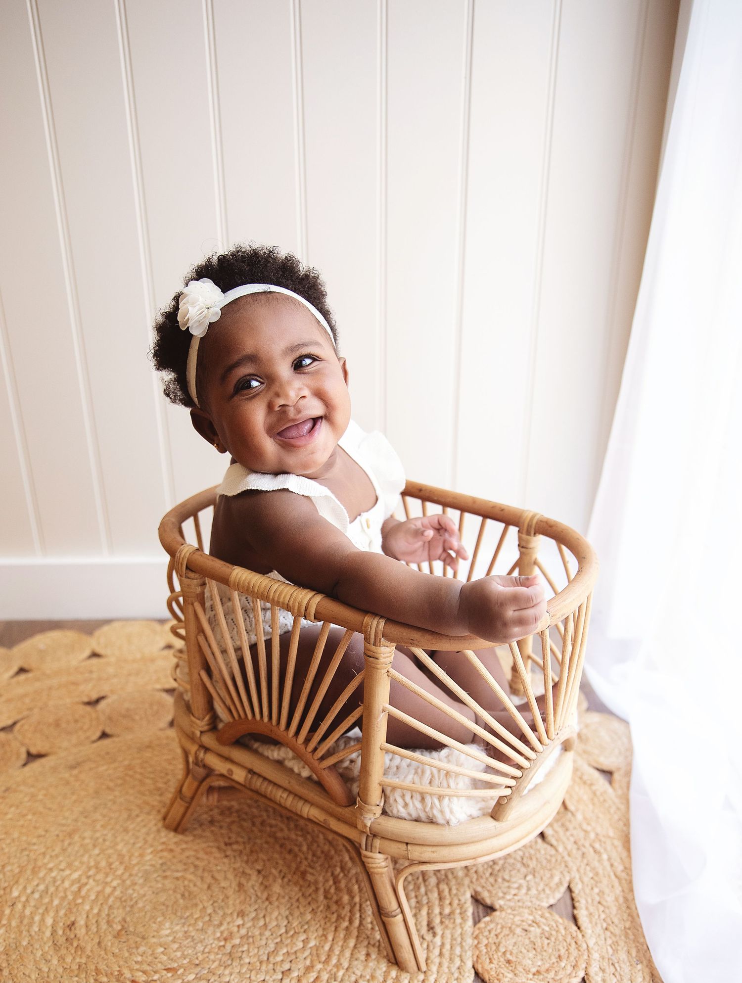 Smiling baby girl in white outfit and floral headband sitting in a rattan chair on a jute rug. During a studio family session in Bexley
