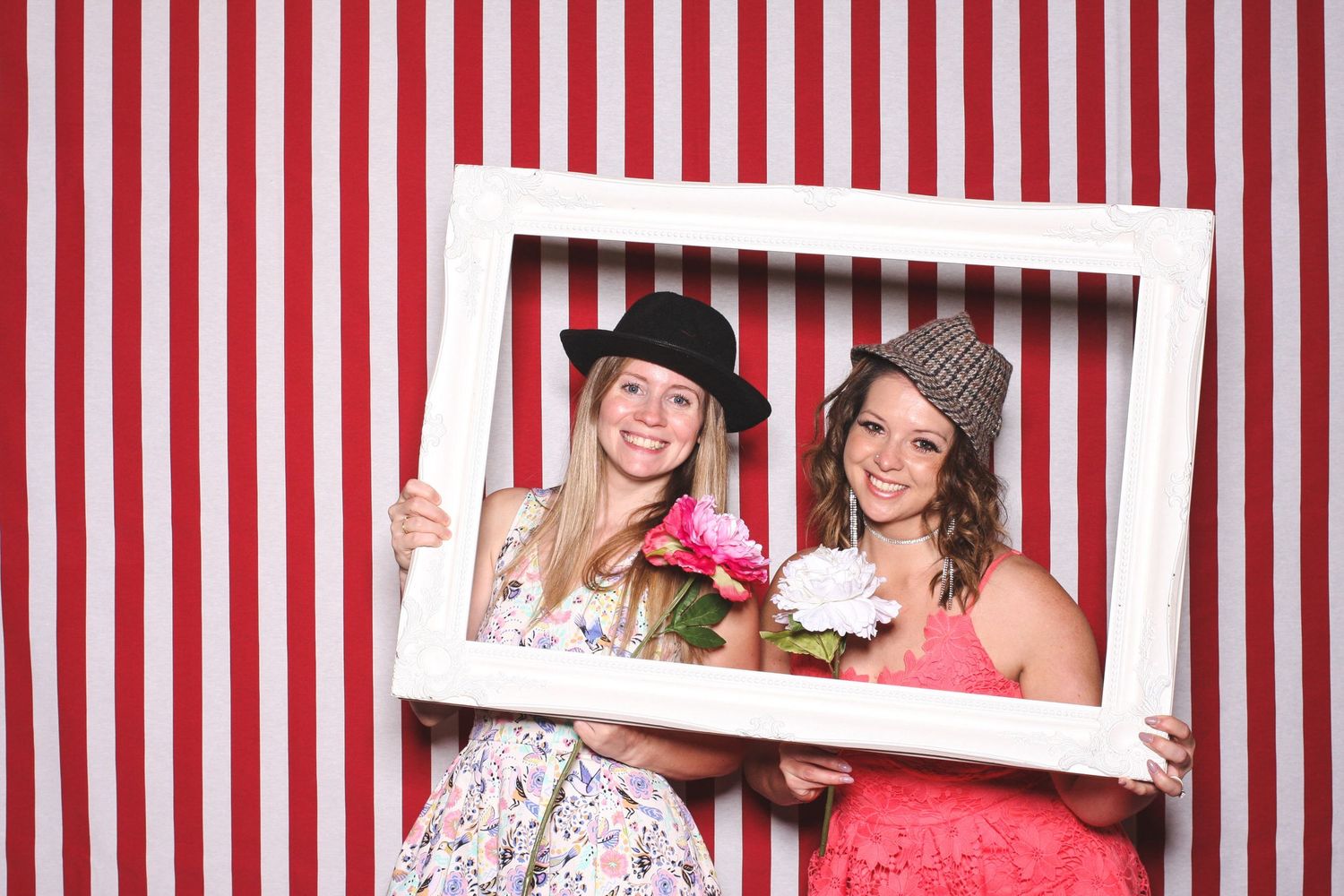 Two smiling women in hats hold a white picture frame and pink flowers against a red and white striped backdrop.