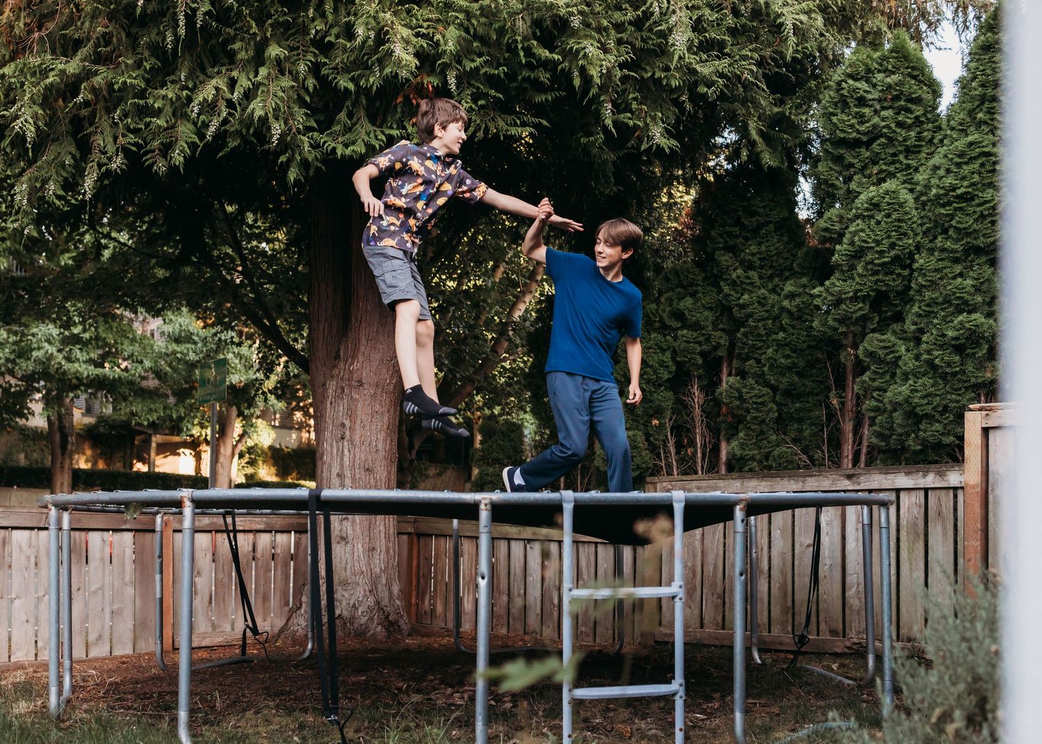 Two boys jumping and holding hands on a backyard trampoline surrounded by trees and wooden fence.