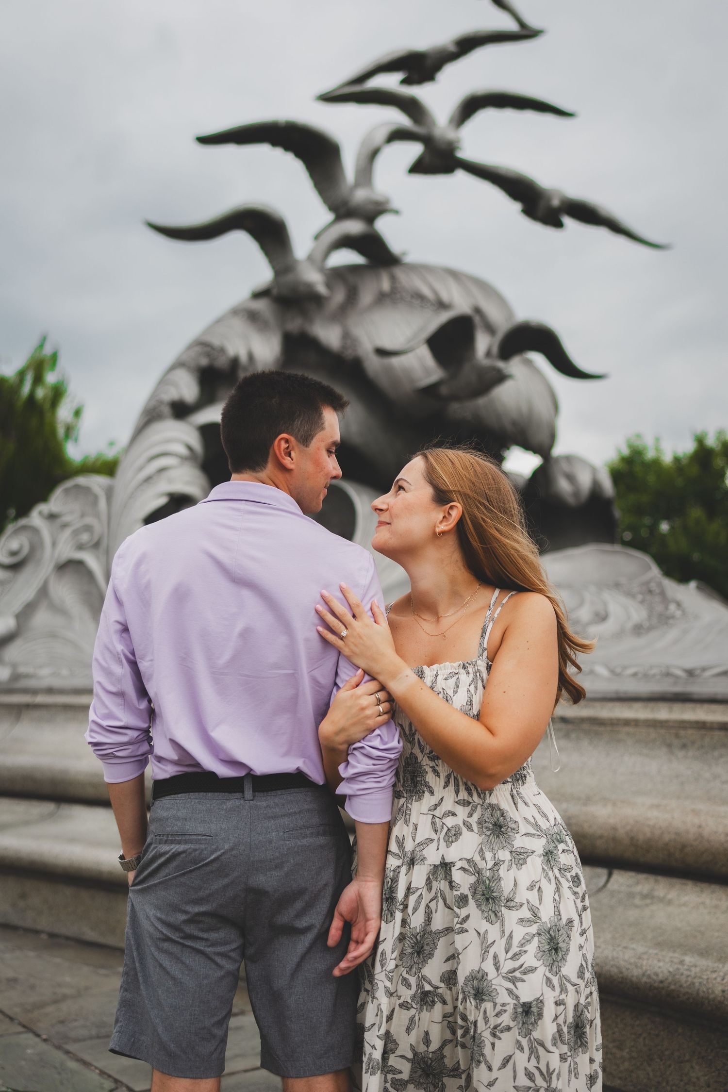 Couple embracing in front of a large bird sculpture at navy merchant marine memorial on a cloudy day after their proposal