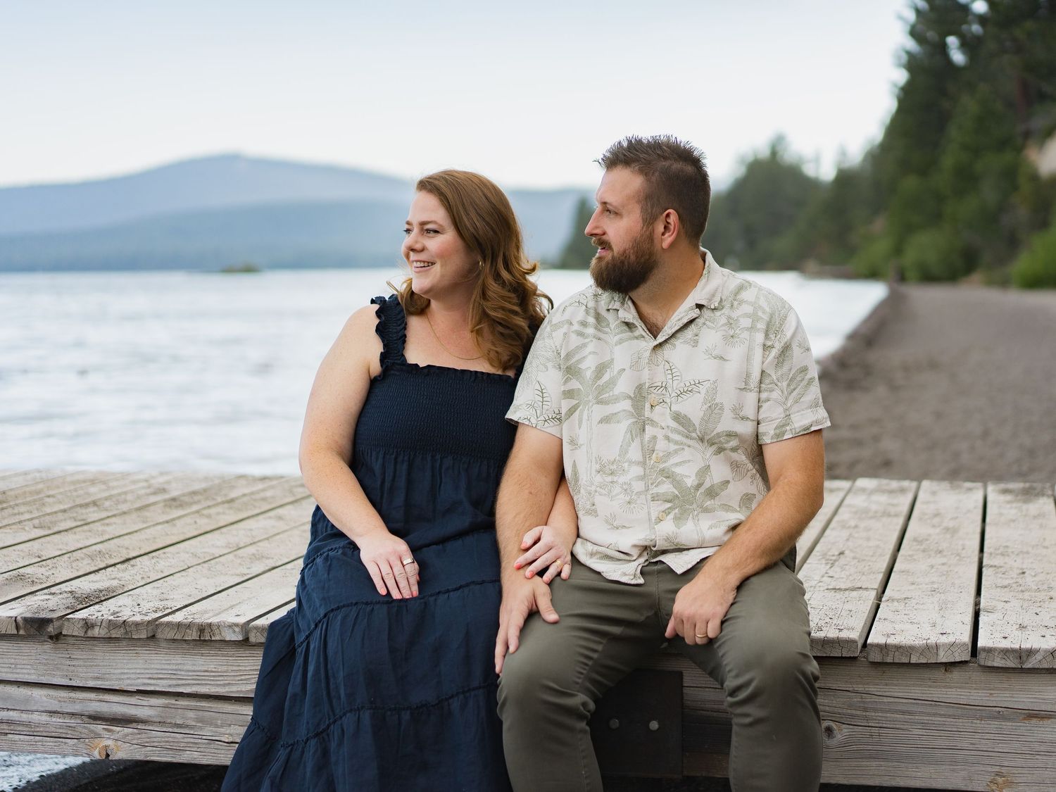 Couple sitting on a wooden dock by a lake with mountain views, smiling together on a peaceful day.