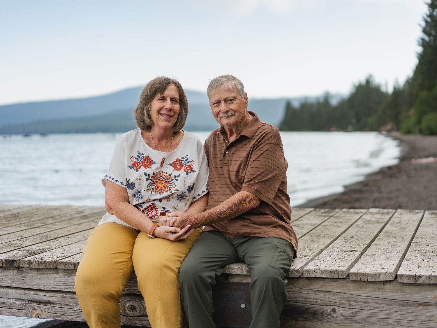 Older couple sitting together on a wooden dock by a lake, smiling and holding hands with mountains in background.