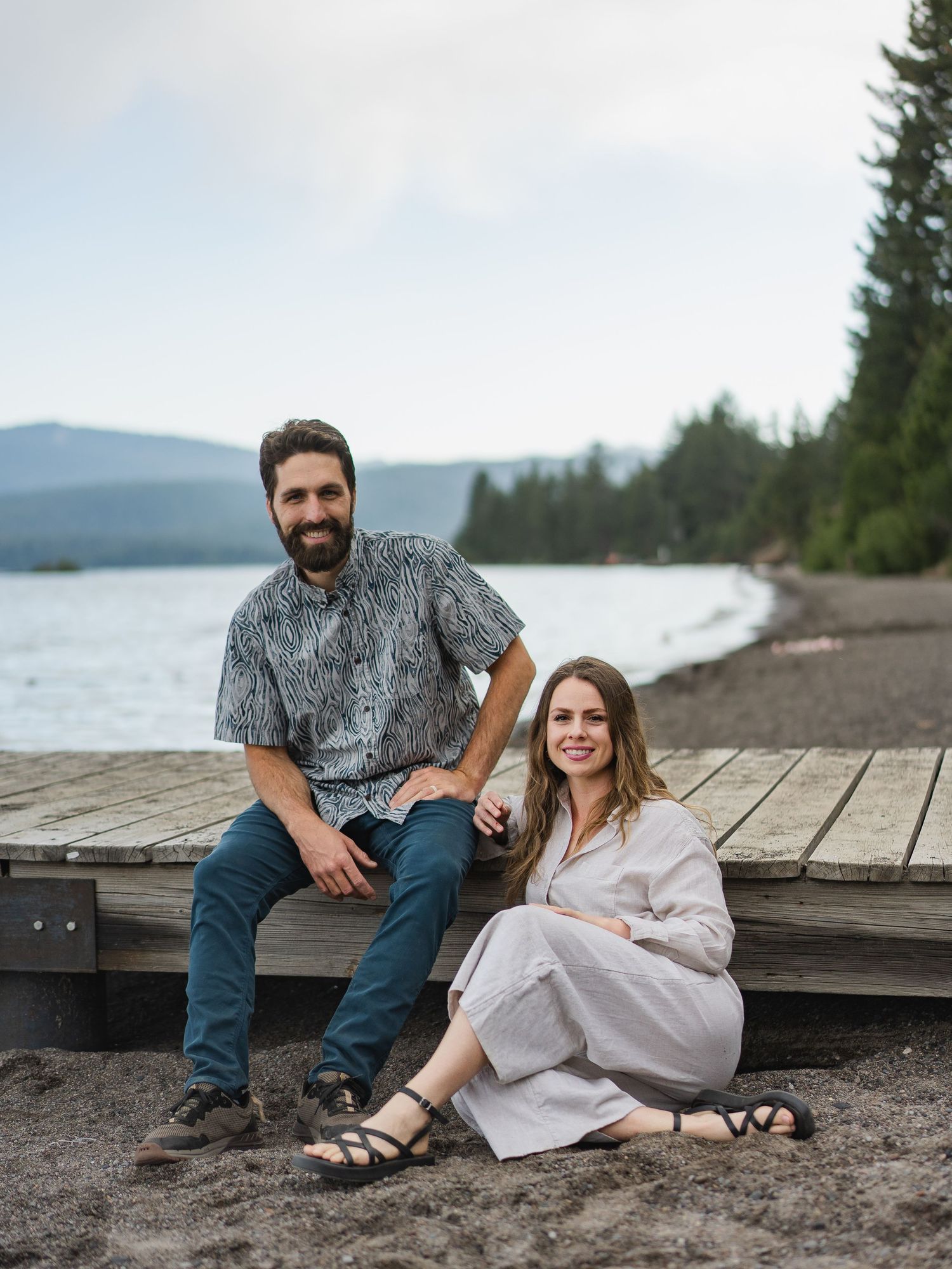 Couple sitting on a wooden dock by a lake surrounded by evergreen trees, smiling outdoors.