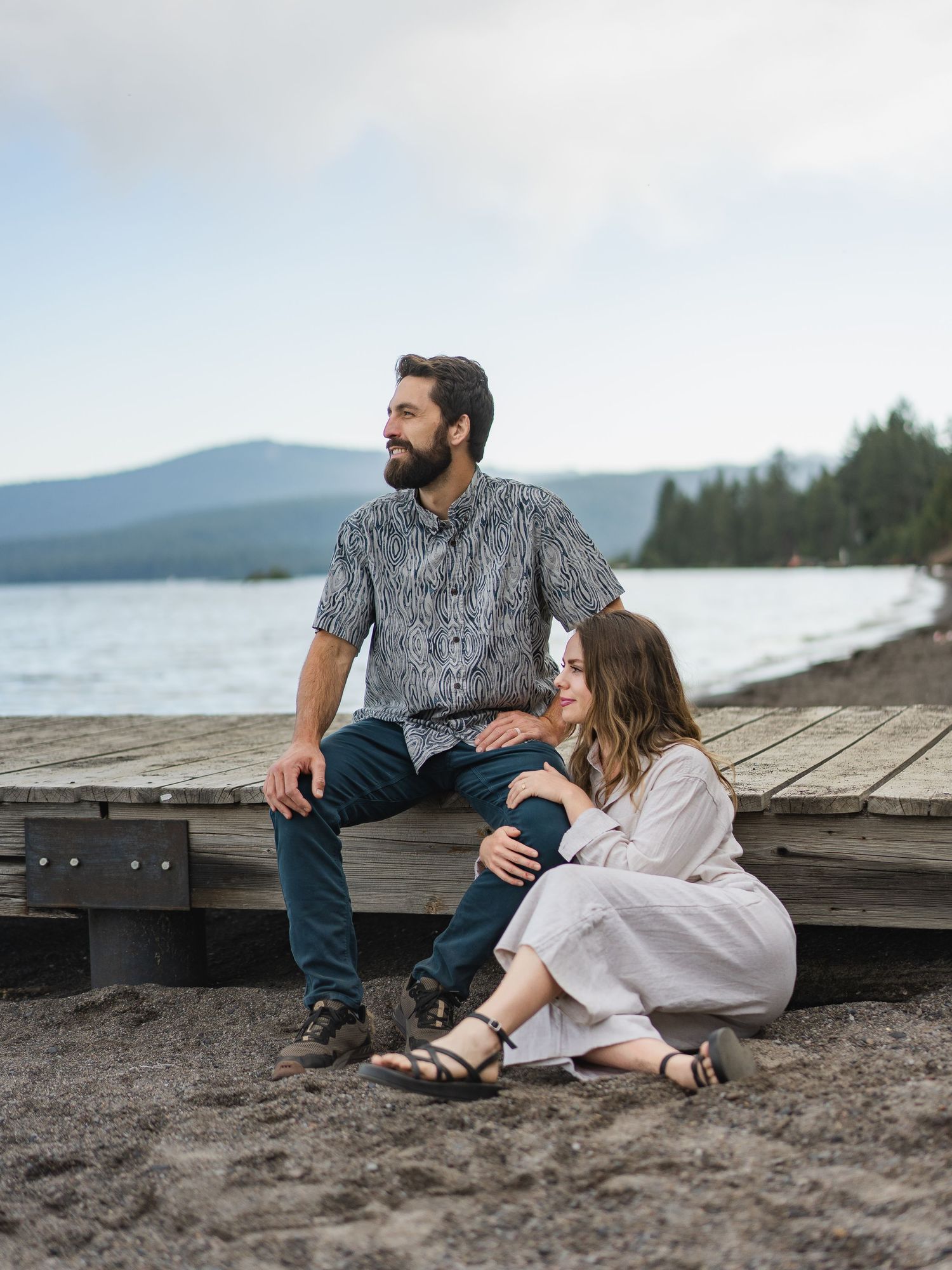 Couple relaxing on a lakeside dock, man seated on edge, woman leaning against him on sandy beach with mountain backdrop.