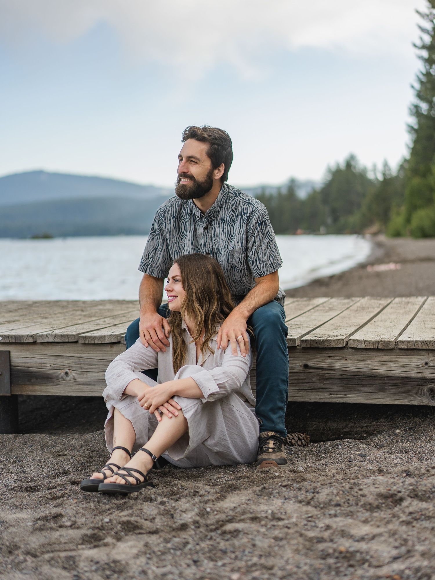 Couple sitting on a lakeside dock, man with beard embracing woman in white dress, mountains in background.