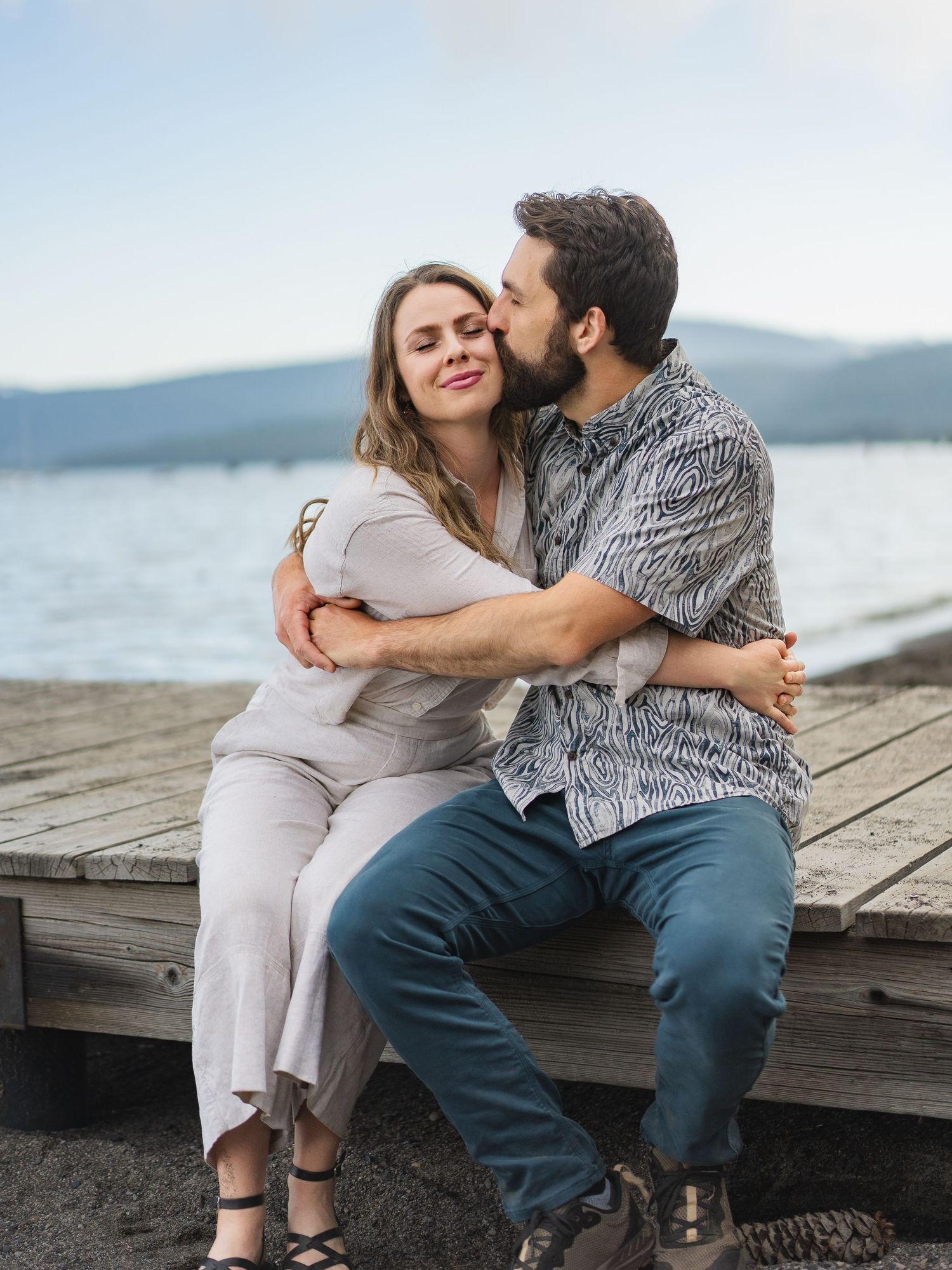 Bearded man kissing woman on cheek while sitting on wooden dock by a scenic mountain lake.