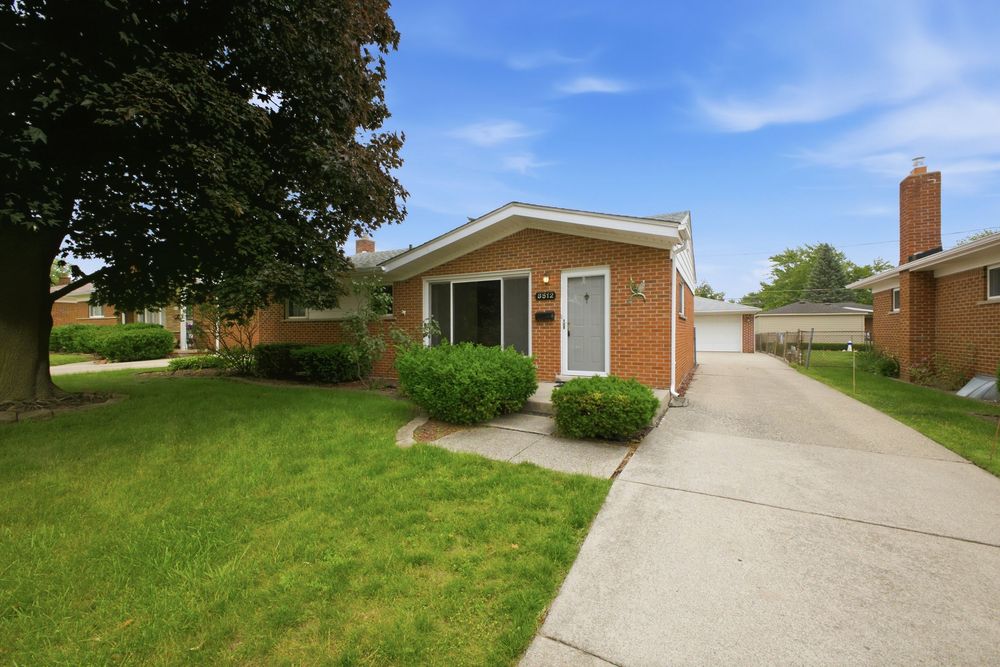 Brick ranch-style home with lush green lawn, mature trees, attached garage, and concrete driveway on a sunny day.