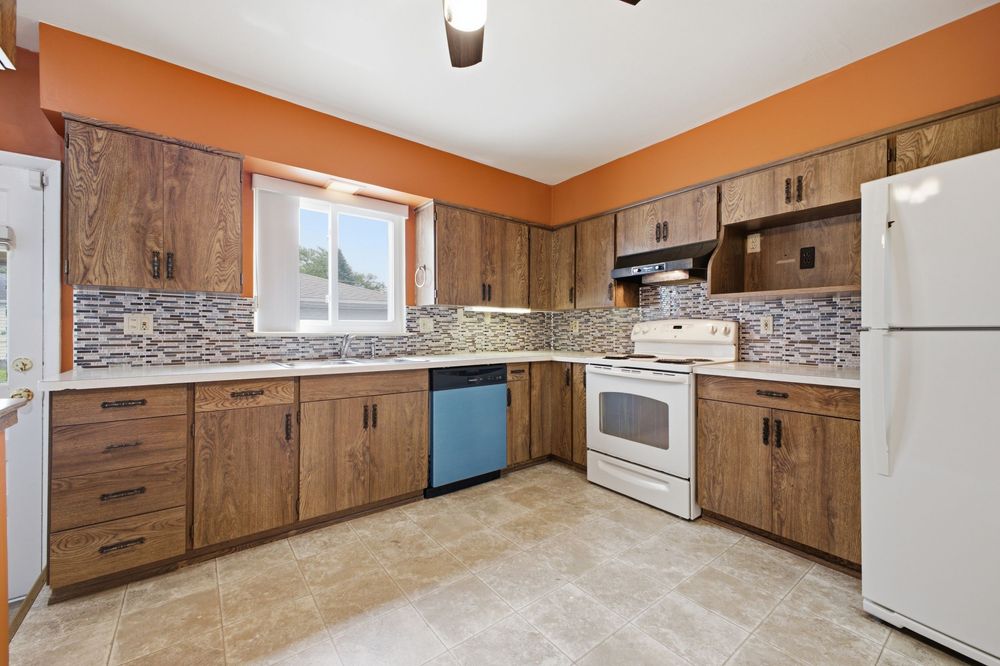Kitchen with brown wood cabinets, mosaic tile backsplash, white appliances, and light tile flooring.