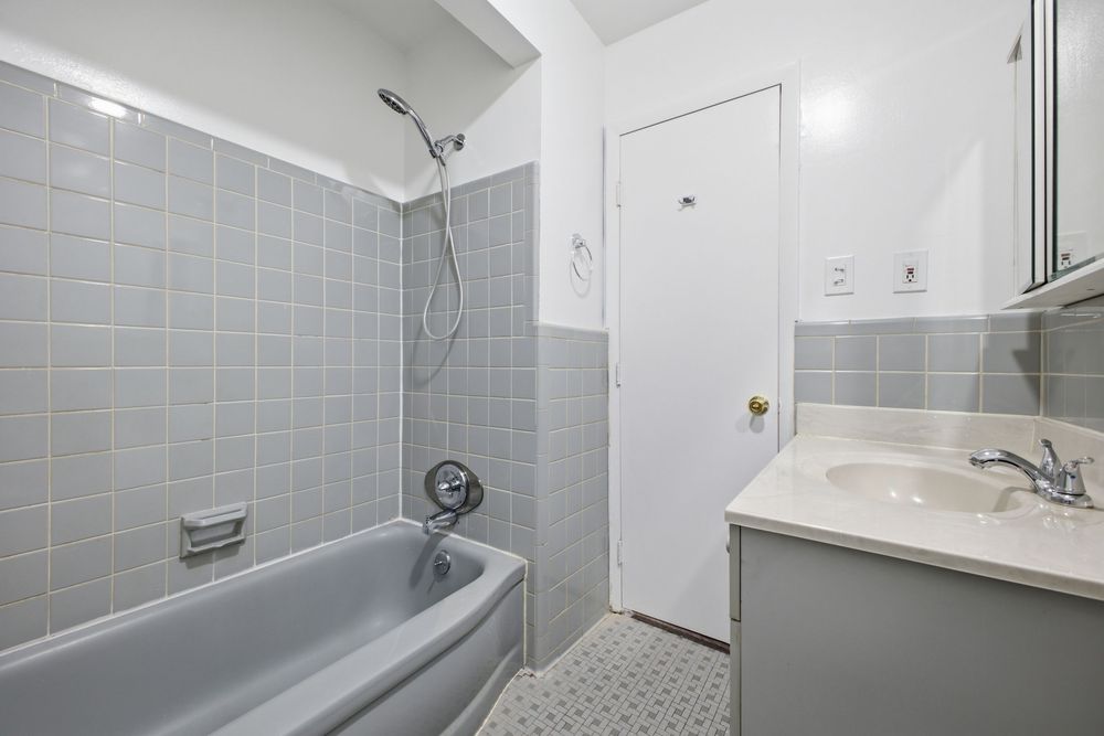 Gray tiled bathroom with bathtub-shower combo, white vanity with sink, mosaic tile floor, and white door.