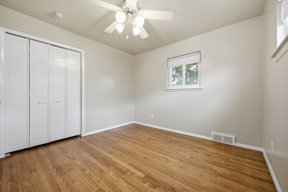 Bright empty bedroom with hardwood floors, white closet doors, ceiling fan, and a window providing natural light.