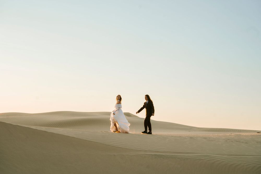Bride and groom hold hands walking across sandy desert dunes at golden hour during an outdoor elopement.