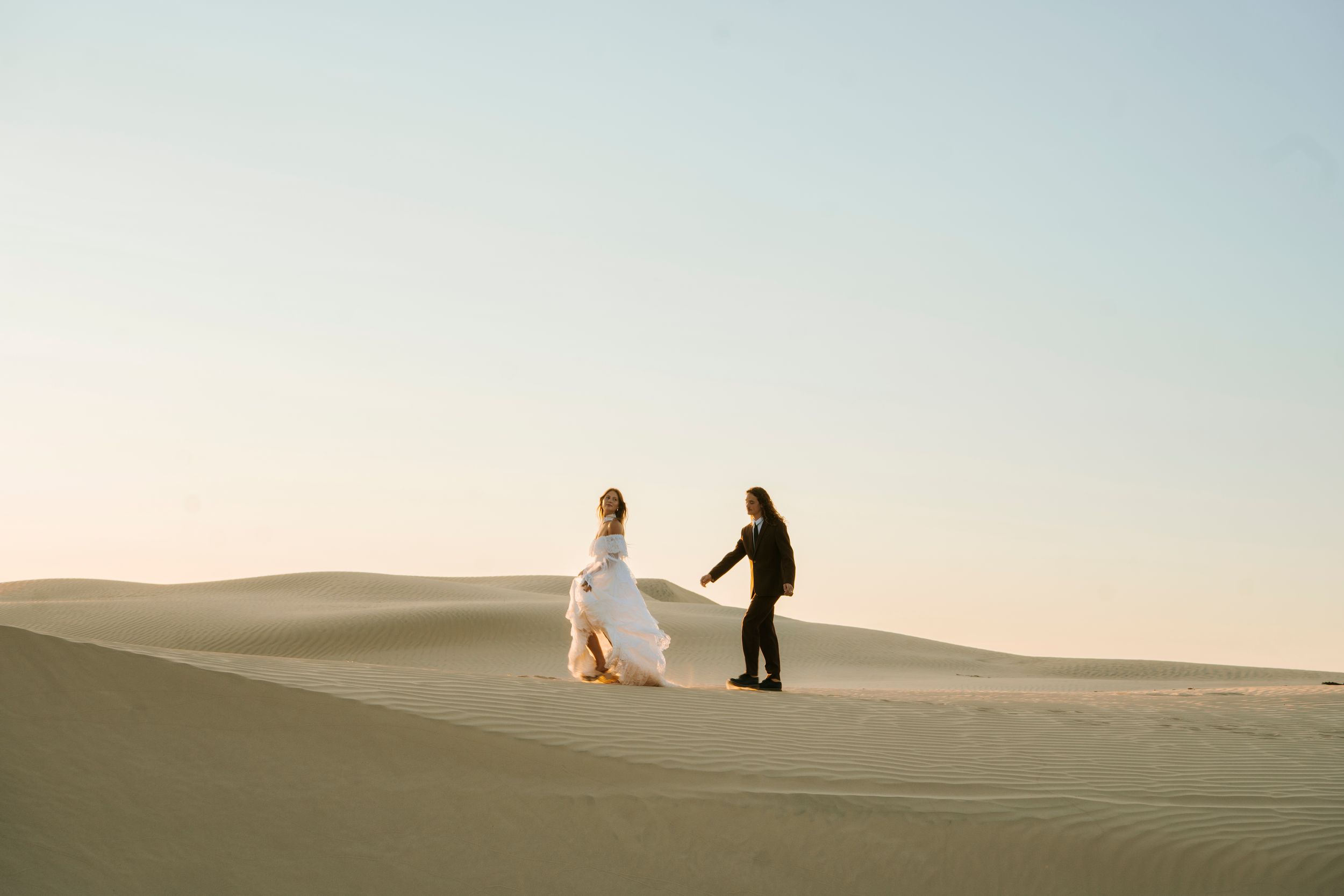 Bride and groom hold hands walking across sandy desert dunes at golden hour during an outdoor elopement.