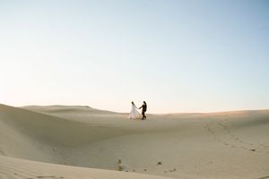 A bride and groom hold hands on vast golden sand dunes under a clear sky in a stunning desert wedding photo.