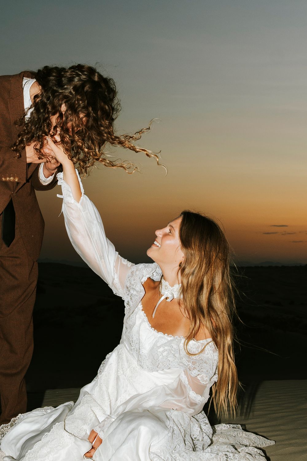 Bride in white lace gown sits on sand dunes, laughing as groom lifts her bouquet at sunset.