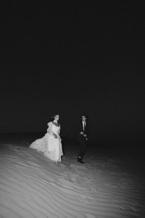 Bride and groom walking on sand dunes at night in black and white wedding photo.