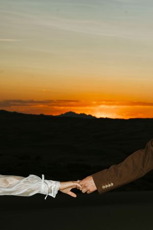 Bride and groom gently touching fingers at sunset with orange sky backdrop.