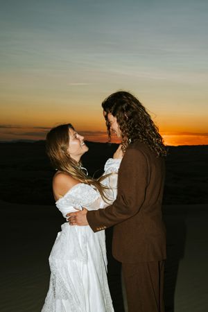 Couple embraces romantically at sunset on sand dunes, bride in white lace gown and groom in brown suit.