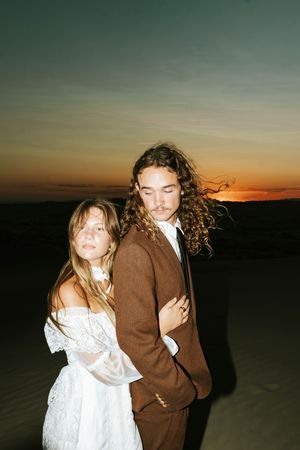 Couple in bohemian wedding attire poses on sand dunes at sunset, woman in white lace dress, man in brown suit.