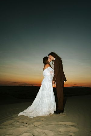 Bride and groom kissing at sunset on sandy dunes, wearing white gown and brown suit.